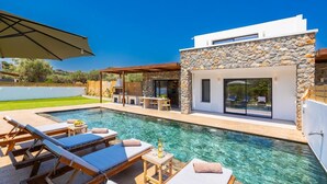 Un área de piscina moderna al aire libre con sillas de descanso, zona de barbacoa y edificio de fachada de piedra.
