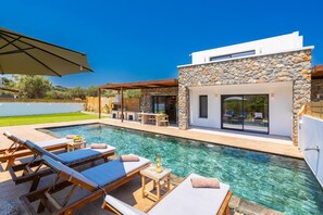 Ein moderner Poolbereich im Freien mit Liegestühlen, einer Grillmöglichkeit und einem Gebäude mit Steinverkleidung.