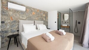 Ett hotellrum med en vägg av sten, en säng med vita och beige sängkläder, ett sängbord, en spegel, en dusch och en handduksställning.