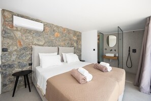 A hotel room with a stone wall, a bed with white and beige bedding, a bedside table, a mirror, a shower area, and a towel rack. - Villa Celestia - Two Bedroom Villa, Sleeps 4 (Afandou)