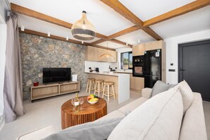 A modern living room with a stone wall, wooden beams, a TV, a dining area, and a kitchen with a black refrigerator. - Villa Celestia - Two Bedroom Villa, Sleeps 4 (Afandou)