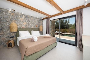A bedroom with a stone wall, a bed with a green headboard, a wooden stool, and a view of a swimming pool through a glass door. - Villa Celestia - Two Bedroom Villa, Sleeps 4 (Afandou)