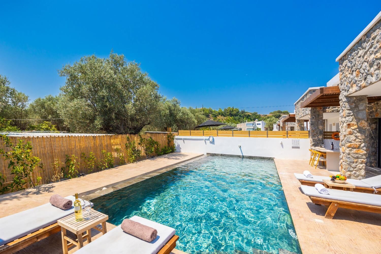 Ein Schwimmbecken mit Liegestühlen und einem Tisch, umgeben von einem Holenzaun und Bäumen.