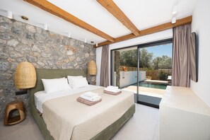 A bedroom with a stone wall, a bed with white linens, a small wooden stool, and a view of an outdoor swimming pool. - Villa Amethysta - Two Bedroom Villa, Sleeps 4 (Afandou)