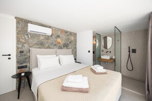 A modern hotel room with a stone accent wall, a bed with white linens, a bedside table, a built-in air conditioning unit, a bathroom with a shower, a mirror, and a towel rack. - Villa Amethysta - Two Bedroom Villa, Sleeps 4 (Afandou)