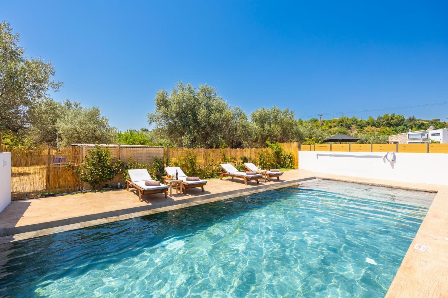 A swimming pool with clear water, surrounded by lounge chairs and a wooden fence.