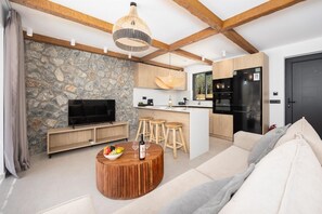 A modern living room with a stone wall, wooden beams, a TV, a round wooden coffee table with fruits and wine glasses, and a sofa. - Villa Amethysta - Two Bedroom Villa, Sleeps 4 (Afandou)