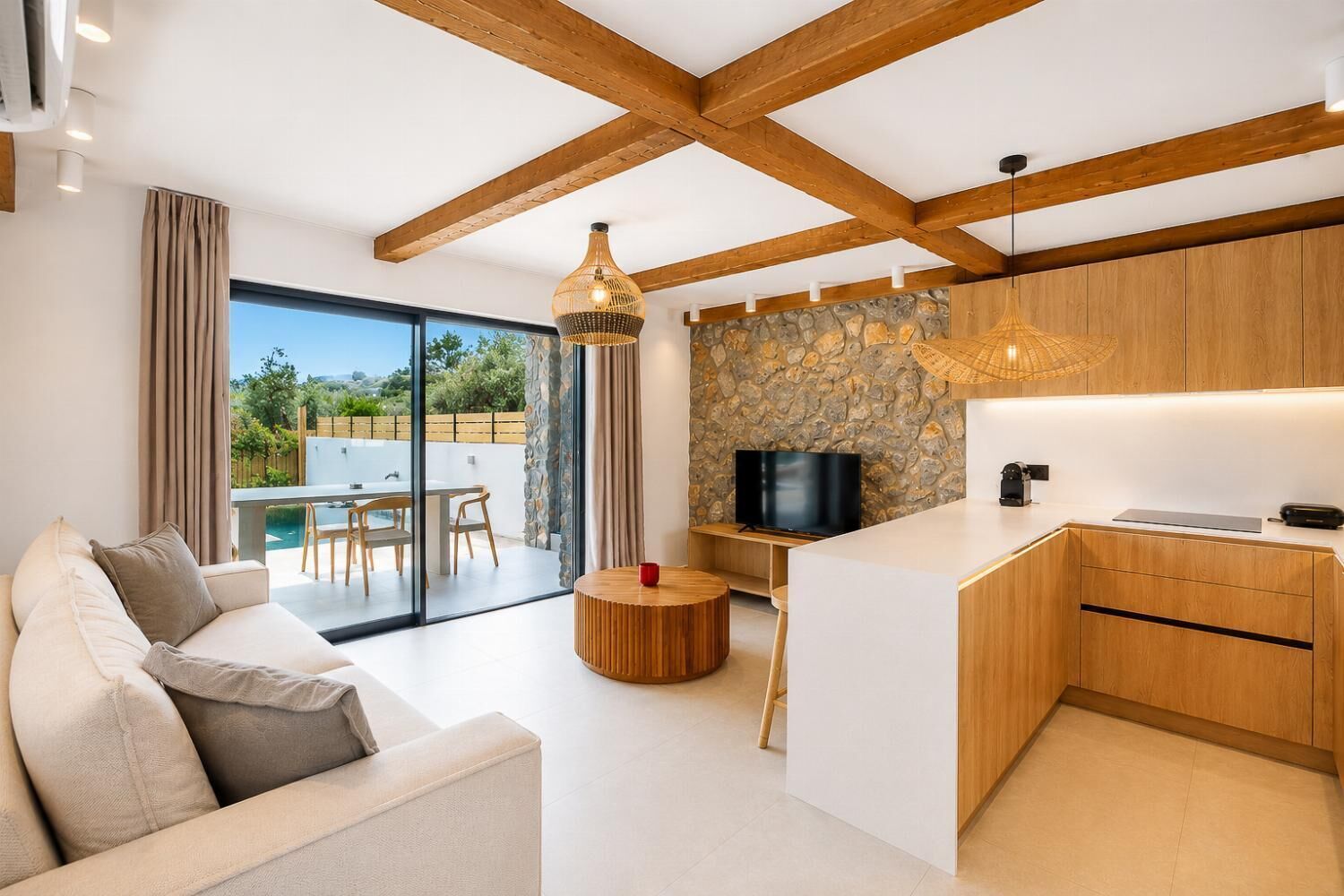 Un salon moderne comprenant un canapé, un coin repas, une cuisine avec un mur de crédence en pierre et une grande fenêtre donnant sur un jardin.