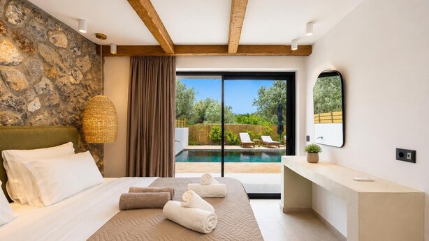 Une chambre avec un mur en pierre, un lit recouvert de draps blancs, une grande fenêtre et une vue sur une piscine et un jardin.
