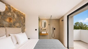Une chambre moderne avec un mur d’accent en pierre, un grand lit avec une literie blanche, un lavabo intégré et une porte en verre donnant sur un balcon.