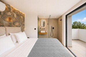 A modern bedroom with a stone accent wall, a large bed with white bedding, a built-in sink, and a glass door leading to a balcony. - Villa Aurelia - Two Bedroom Villa, Sleeps 4 (Afandou)