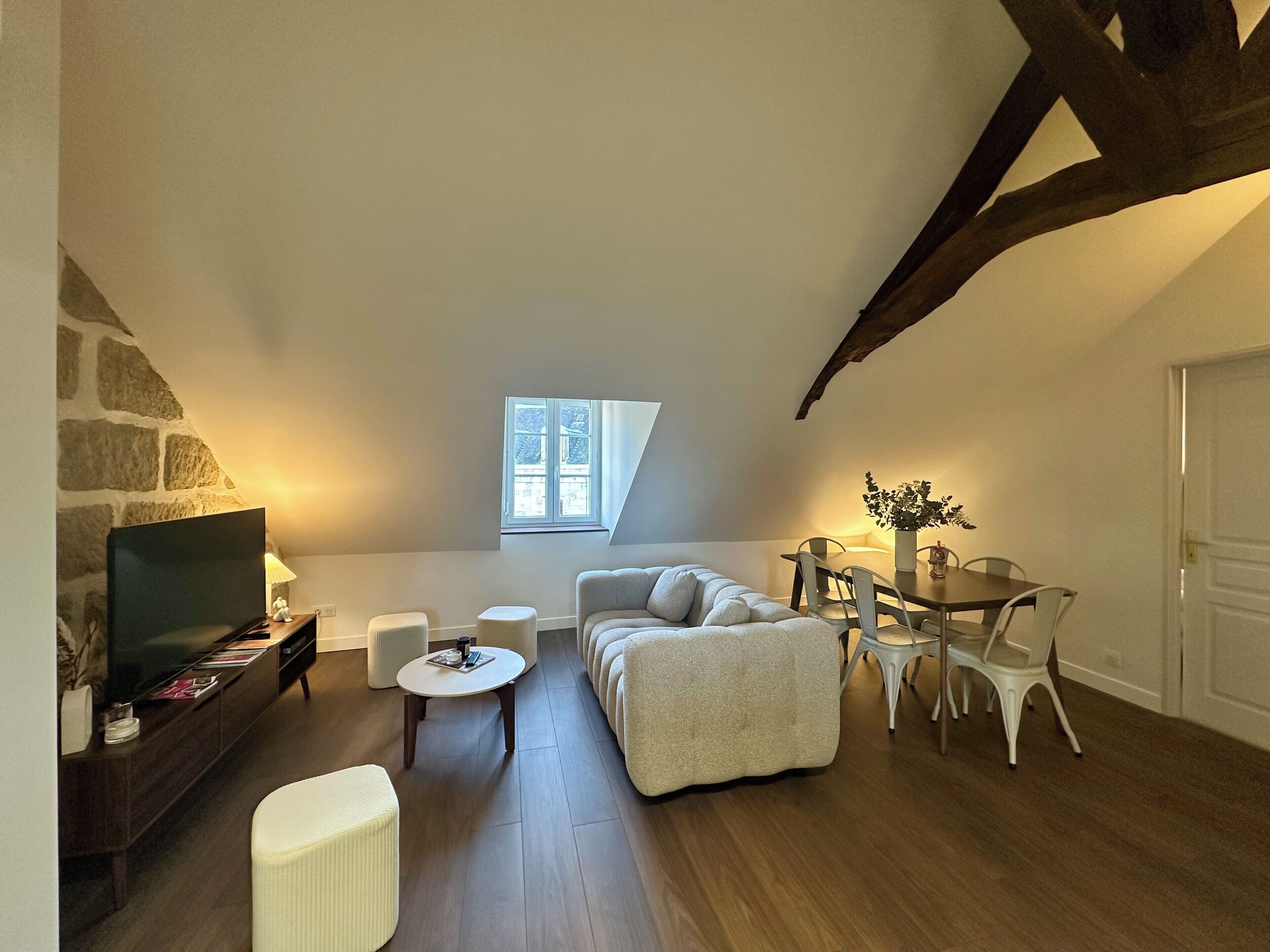 Una sala de estar estilo loft acogedora, con pared de piedra, televisor, sofá, mesa de comedor con sillas y un ventanal pequeño.