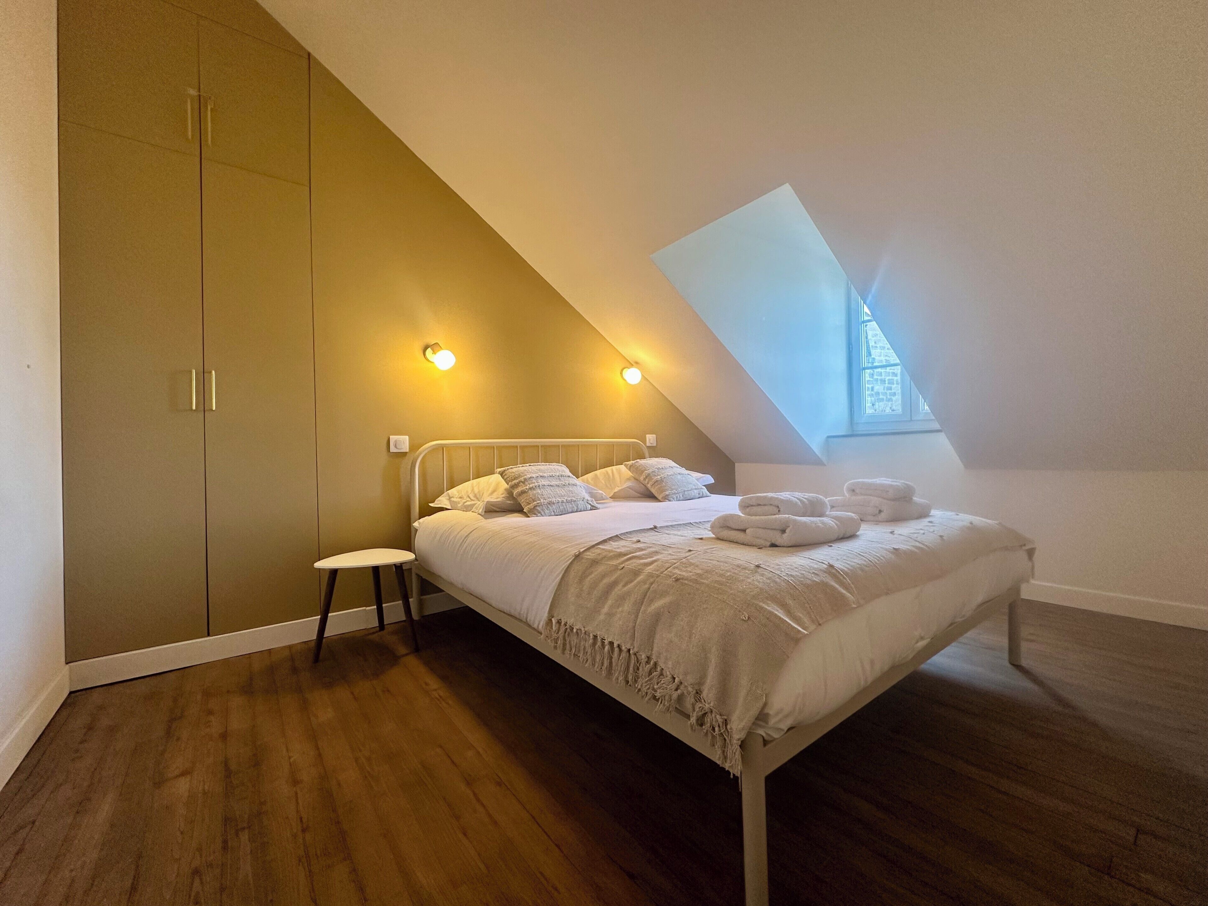 Une chambre à coucher comprenant un lit, un petit tabouret, une armoire et un plafond en pente avec une lucarne.