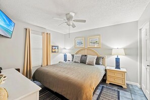 A bedroom with a bed, two bedside tables with lamps, a ceiling fan, a TV on the wall, and a window with blinds. - Pelican Beach Resort · Ocean Views | 3 Pools, Hot Tub, Courts, Gym & Balcony (Destin)