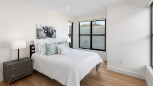 A bedroom with a bed, a nightstand, a lamp, a window with a view, and a wall-mounted artwork.