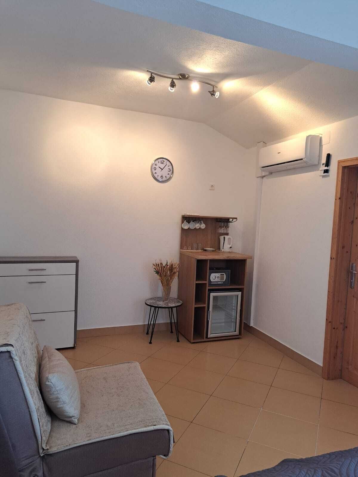 A compact living space with a sofa, a small table with a vase, a clock, a mini fridge, and a wooden cabinet.