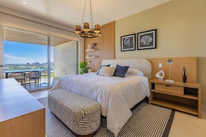 A modern bedroom with a large bed, a desk, and a balcony with a view of the desert. - Vista Del Arco - Vista Vela Sunset (Cabo San Lucas)
