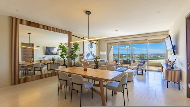 A modern dining area with a large wooden table and chairs, a TV, and a view of the city through large windows. - Vista Del Arco - Vista Vela Sunset (Cabo San Lucas)