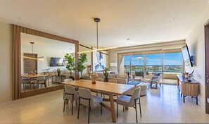 A modern dining area with a large wooden table and chairs, a TV, and a view of the city through large windows. - Vista Del Arco - Vista Vela Sunset (Cabo San Lucas)