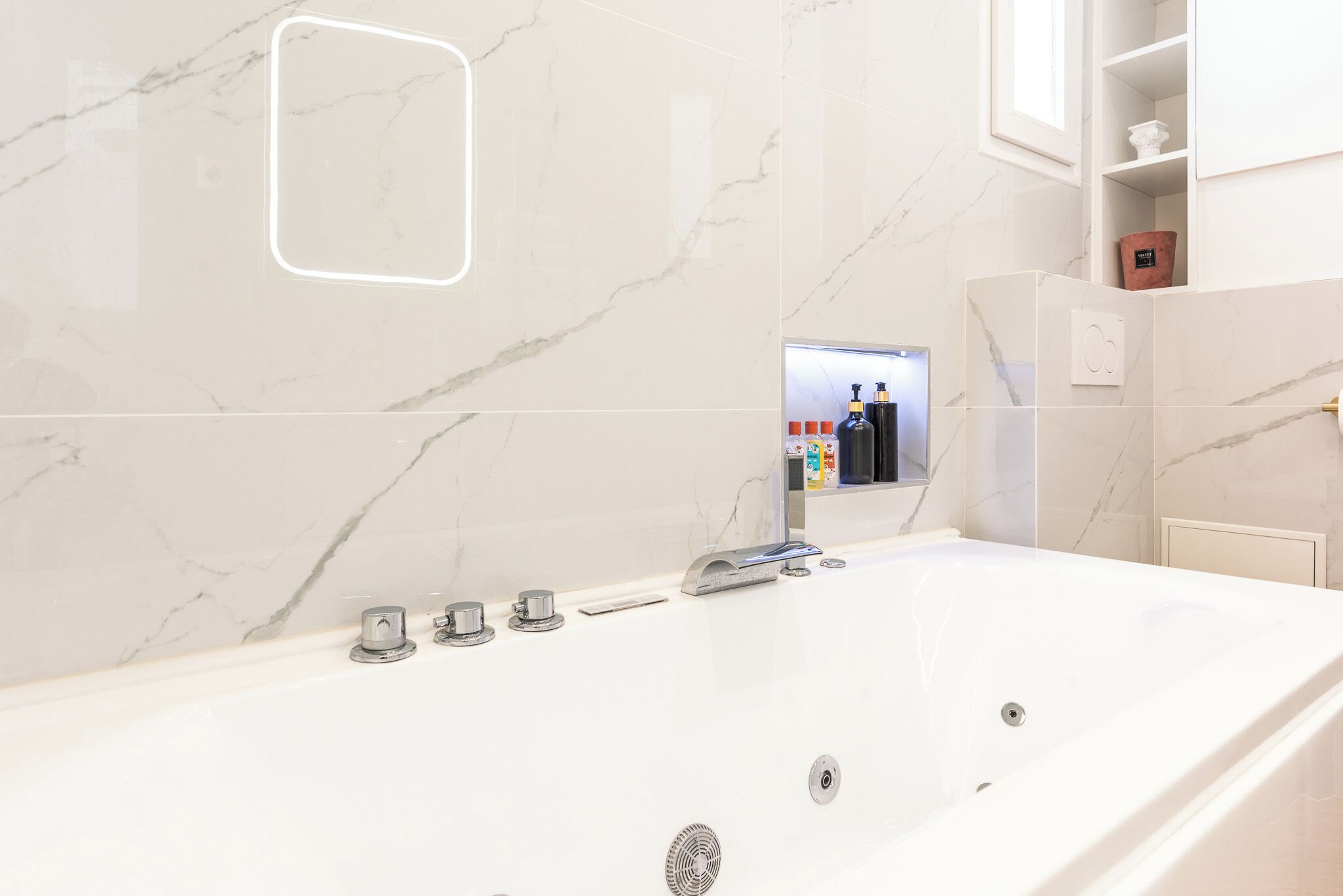 Une salle de bain moderne avec un mur en marbre, une baignoire blanche et une petite étagère contenant des articles de toilette.