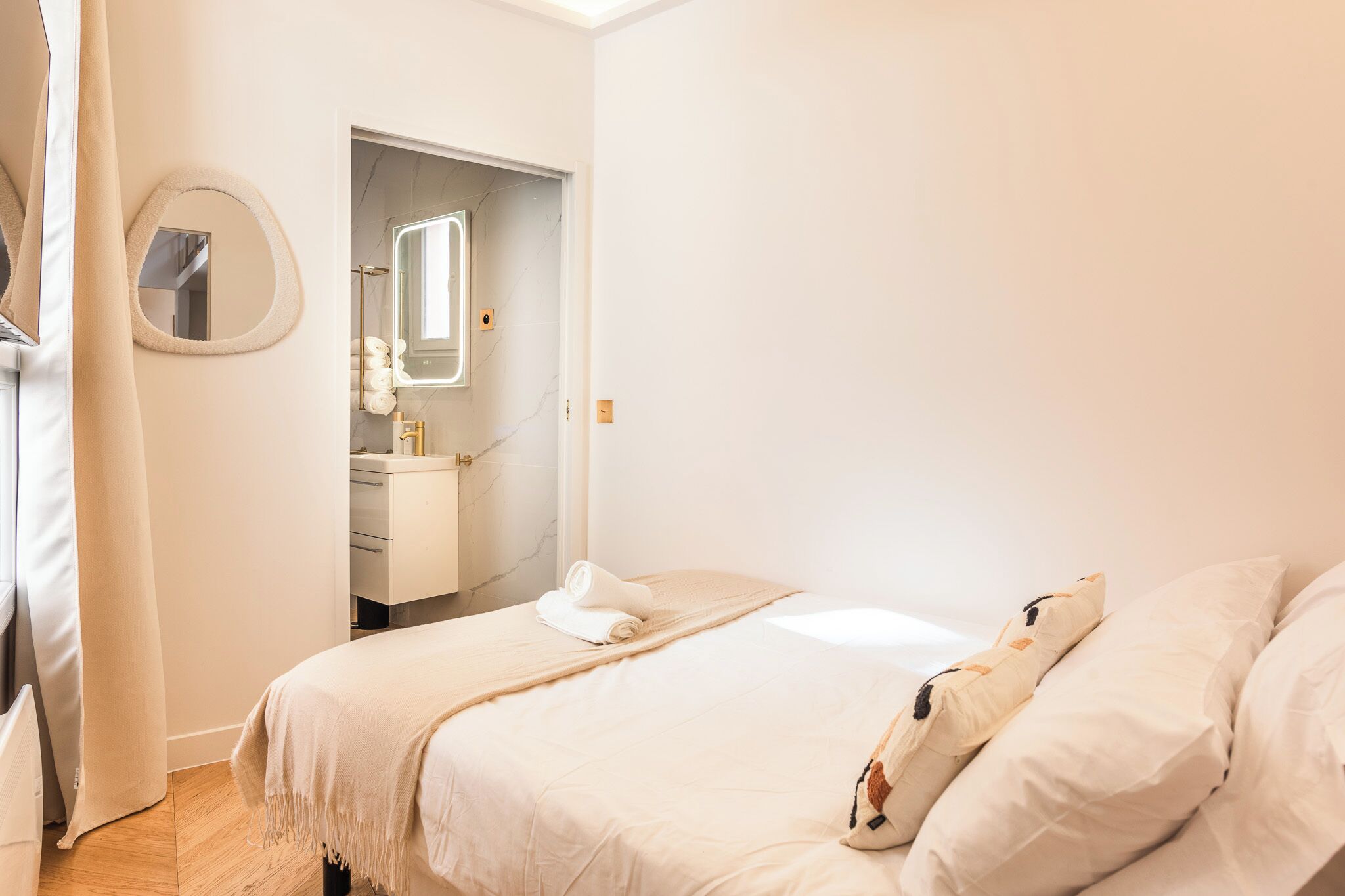 Un dormitorio con cama, espejo redondo y baño visible a través de una puerta abierta.