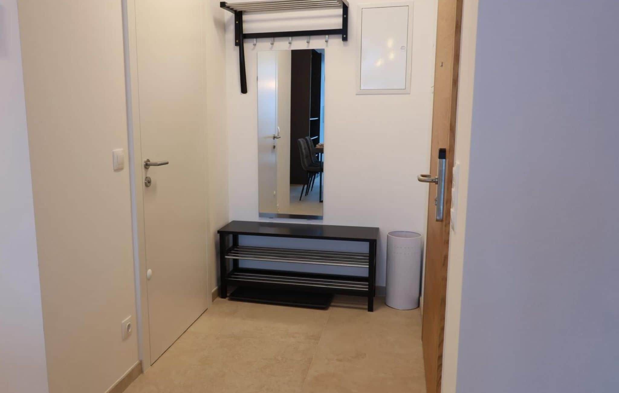 A narrow hallway with a door on the left, a bench with a towel rack in the center, and a trash can on the right.