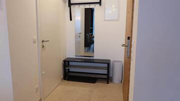 A narrow hallway with a door on the left, a bench with a towel rack in the center, and a trash can on the right.