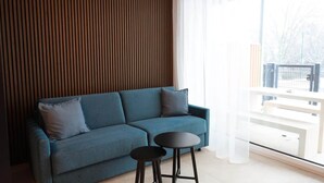A modern living room with a blue sofa, two black stools, and a large window with sheer curtains. - Apartments Kellergasse - Blue Apartment (Purbach am Neusiedlersee)