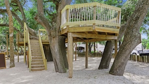 Una estructura de casa en árbol de madera con una escalera y una plataforma, rodeada de árboles y suelo arenoso.