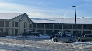 Un edificio con techo nevado y varios balcones. Hay varios coches aparcados delante, y el suelo está parcialmente cubierto de nieve.