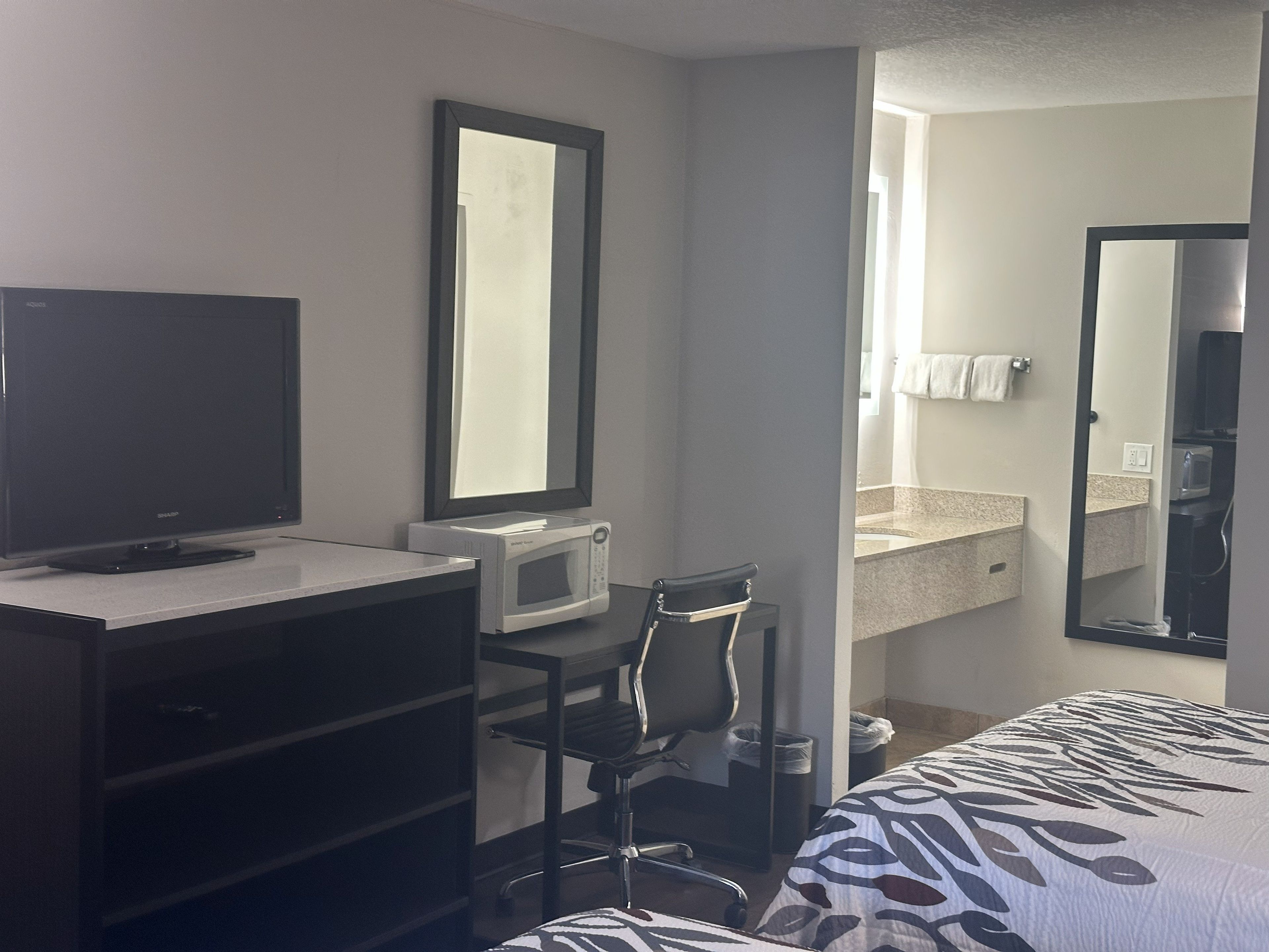 A hotel room with a bed, a desk with a computer, a TV, a microwave, a chair, and a bathroom visible through an open door.