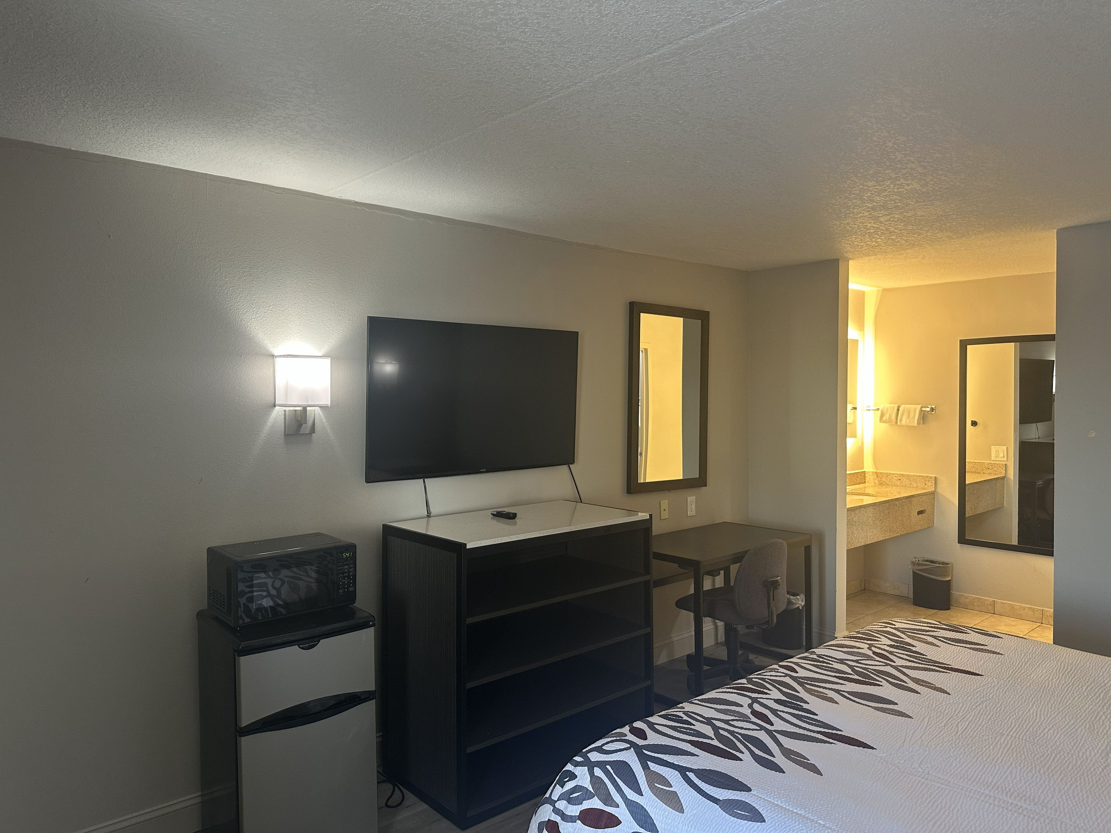Ein Hotelzimmer mit Flachbildfernseher, Schreibtisch, Stuhl, einem Bett mit gemustertem Überwurf und Spiegel. Ein Badezimmer ist durch eine geöffnete Tür erkennbar.
