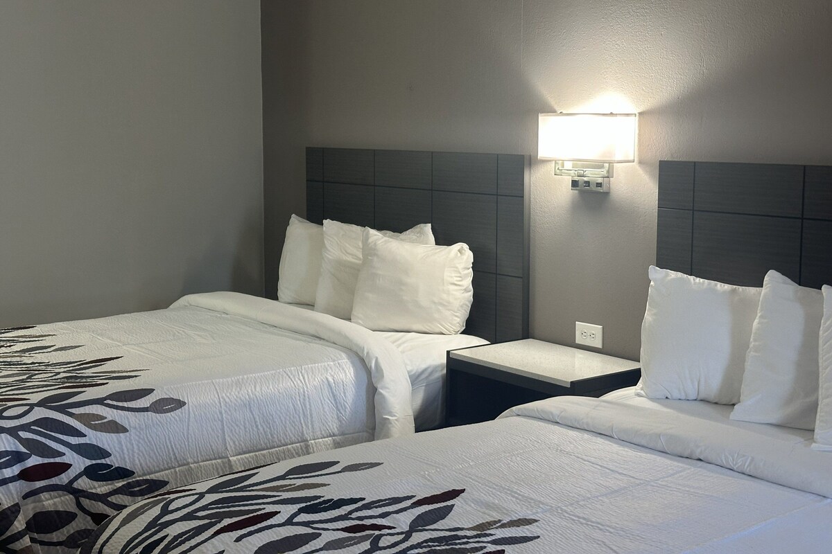Habitación de hotel con dos camas, cada una con ropa de cama blanca y edredón estampado con motivos de hojas. Sobre las camas, hay una lámpara de pared.
