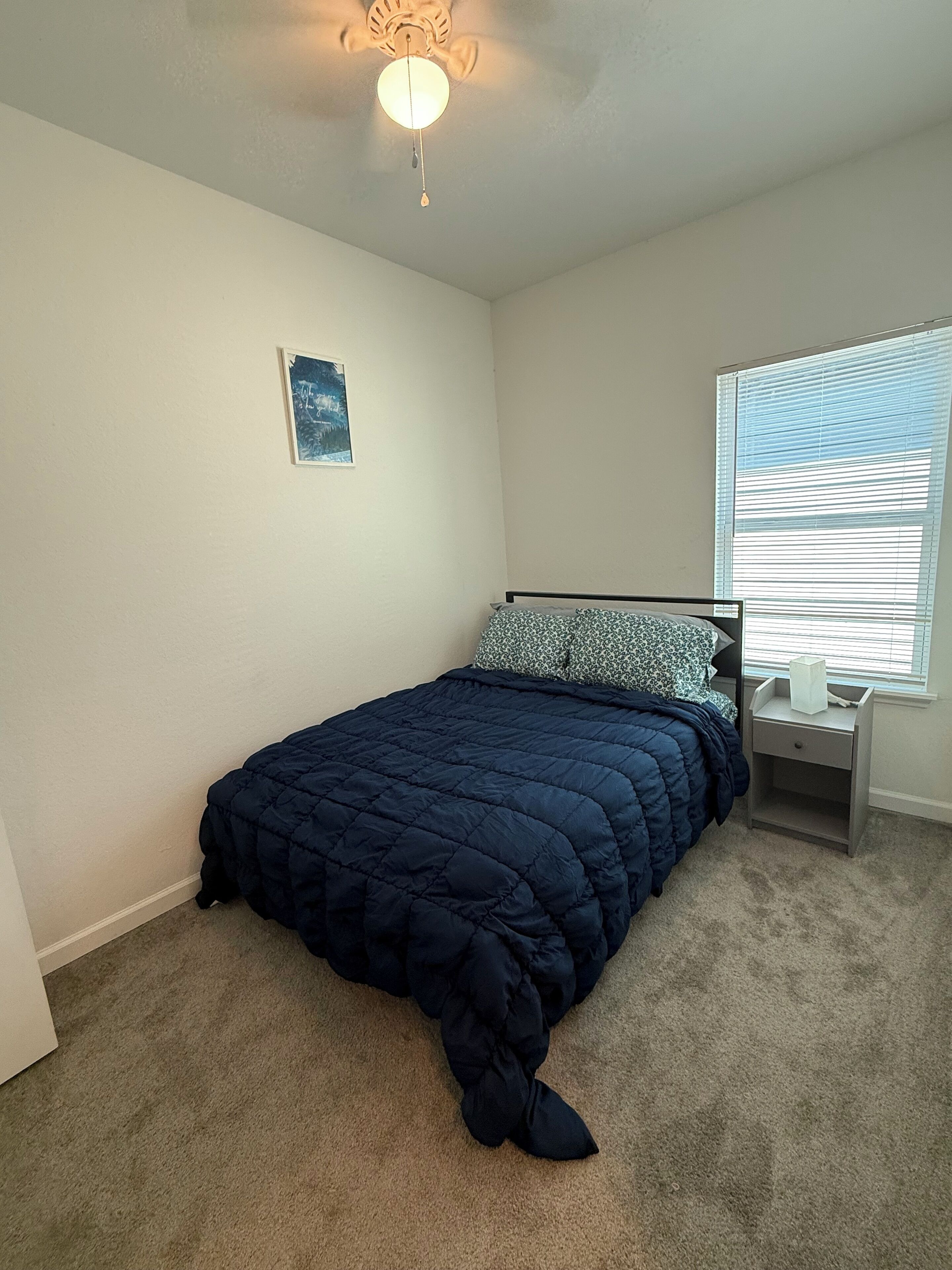 Un dormitorio con cama, mesita de noche, ventilador de techo, ventana con persianas e imagen colgada en la pared.