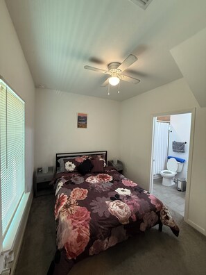 Een slaapkamer met een bed, nachtkastje, plafondventilator, raam met zonwering en een deur naar een badkamer.