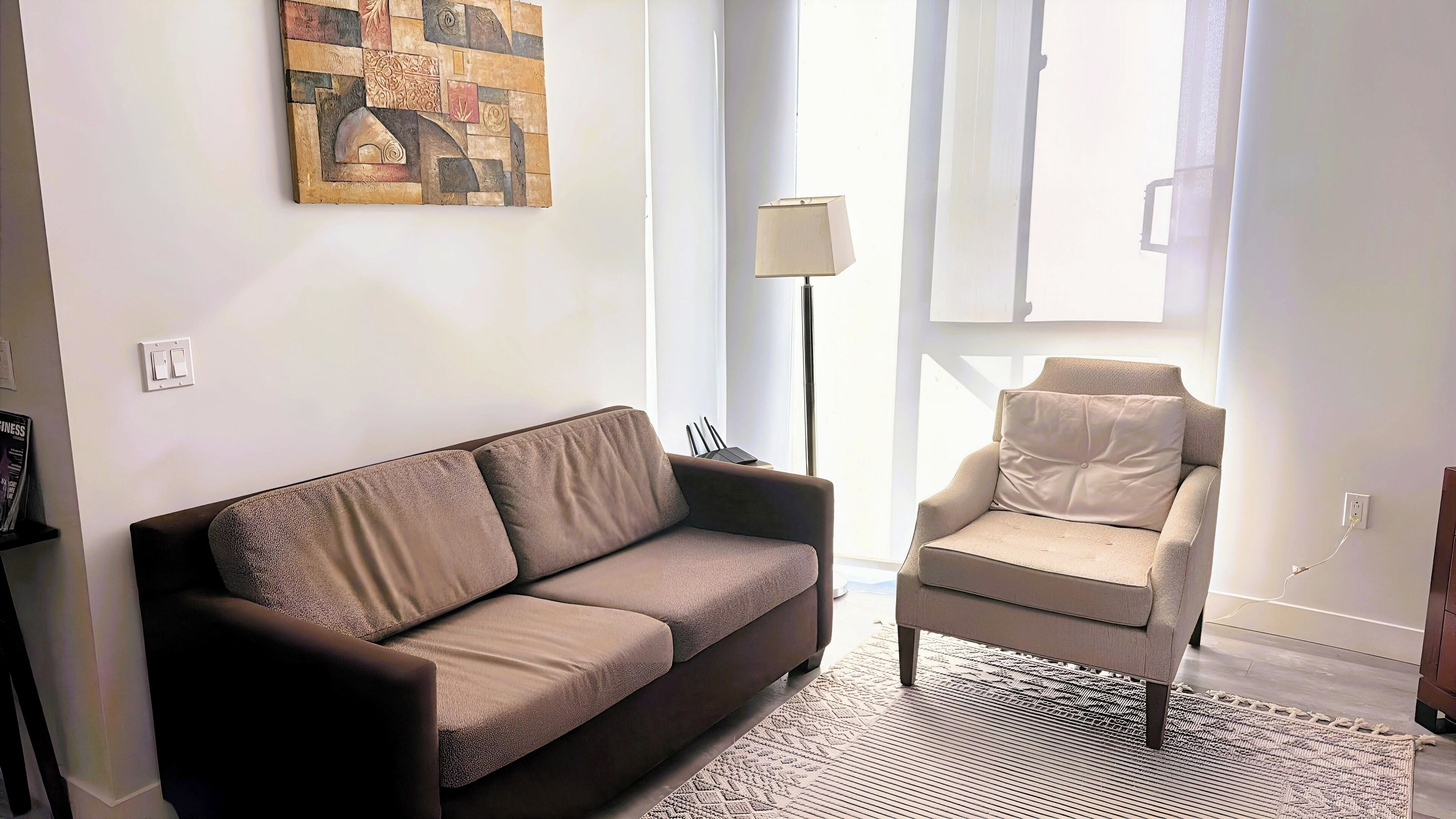 Een woonkamer met een bruine bank, een beige fauteuil, een vloerlamp en een schilderij aan de muur.