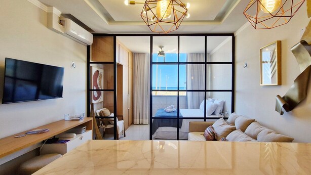 A modern hotel room with a flat-screen TV, a dining table, a sofa, a balcony with a view, and geometric wall art. - COPIU0100 - Edifício Corsário (Salvador)