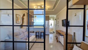 A modern living room with a sofa, a coffee table, and a TV. There is a kitchen area with a refrigerator and a dining table. The room has a glass partition with a view of the beach. - COPIU0100 - Edifício Corsário (Salvador)