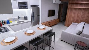 A modern kitchen with a central island, a dining area with bar stools, a sofa, and a TV mounted on the wall. - Apartamento novo de alto padrão 223NW (Brasilia)