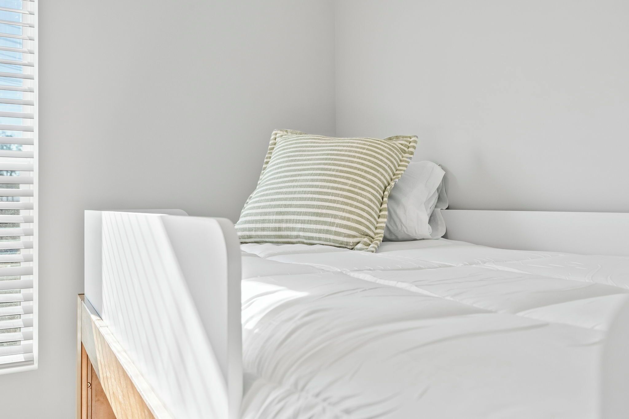 Una cama bien tendida con ropa blanca y una almohada a rayas en una habitación sencilla y despejada, con paredes blancas y una ventana con persianas.