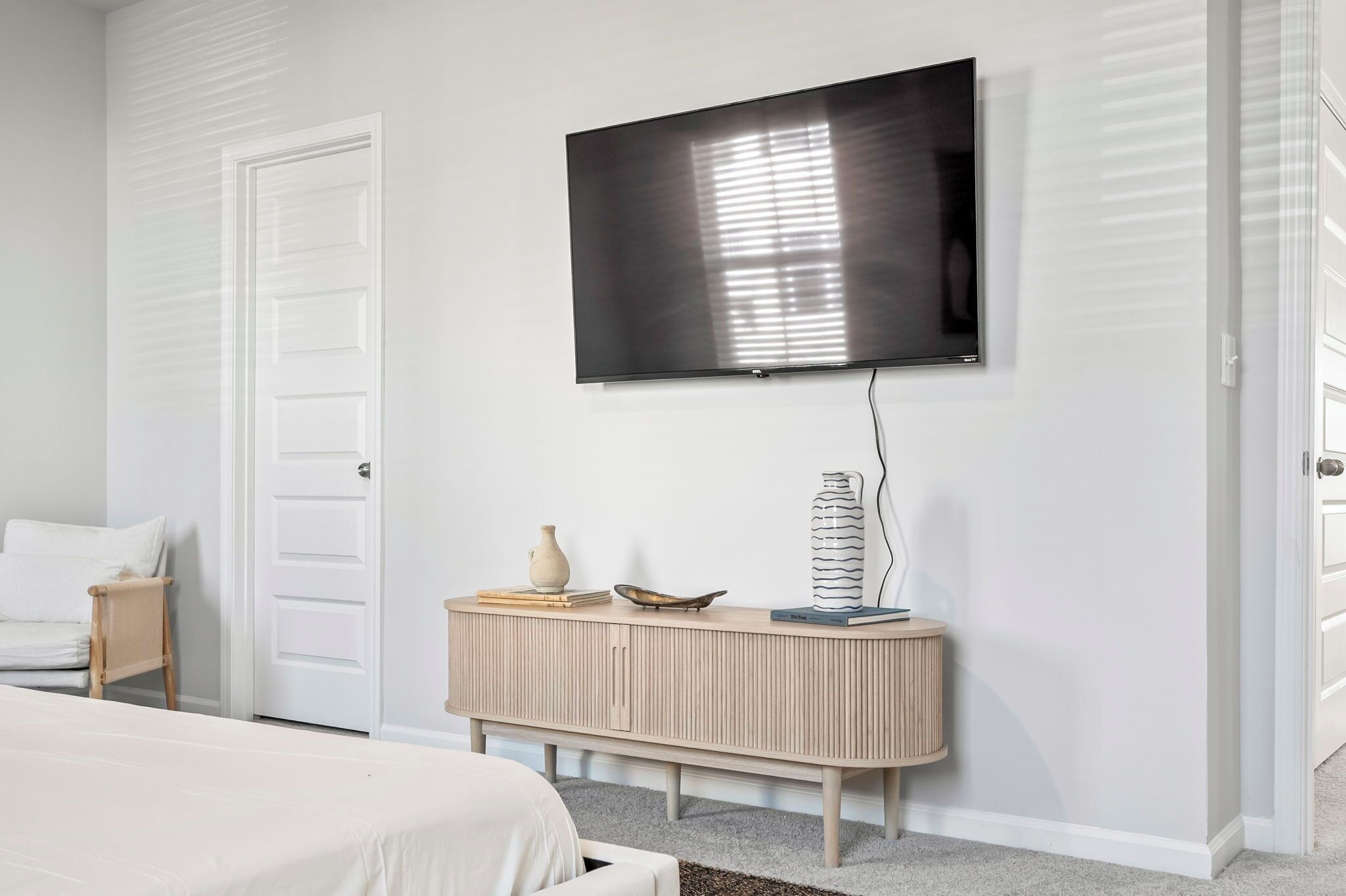 A bedroom with a bed, a bedside table with a lamp, a TV mounted on the wall, and a console table with decorative items.