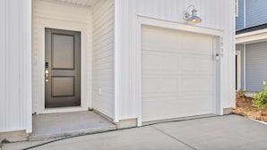Une maison moderne à deux étages, avec une façade blanche, une porte d’entrée sombre et un garage surmonté d’un luminaire.