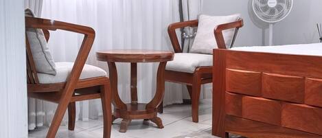 A room with a wooden bed, two wooden chairs, a small round table, and a standing fan.