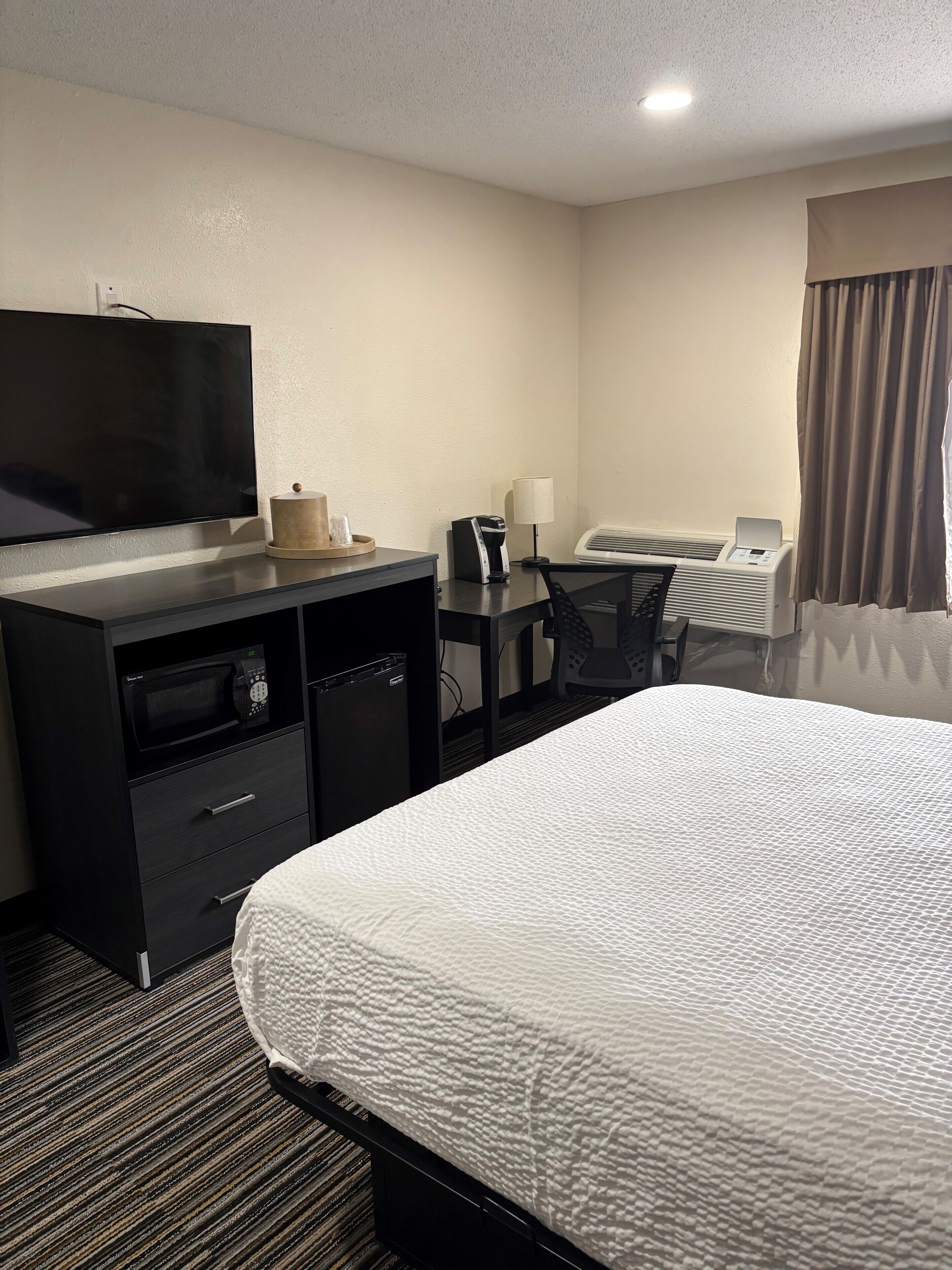 Quarto de hotel com cama, escrivaninha com computador, televisão, cafeteira e ar condicionado.