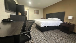 Habitación de hotel con cama, escritorio con teléfono, silla, mesita de noche con lámpara y televisor montado en la pared.