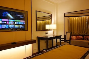 A hotel room with a bed, a desk, a TV displaying a hotel app, a mirror, and a window with curtains. - Ramada By Wyndham Jaisalmer Barmer Road (Jaisalmer)