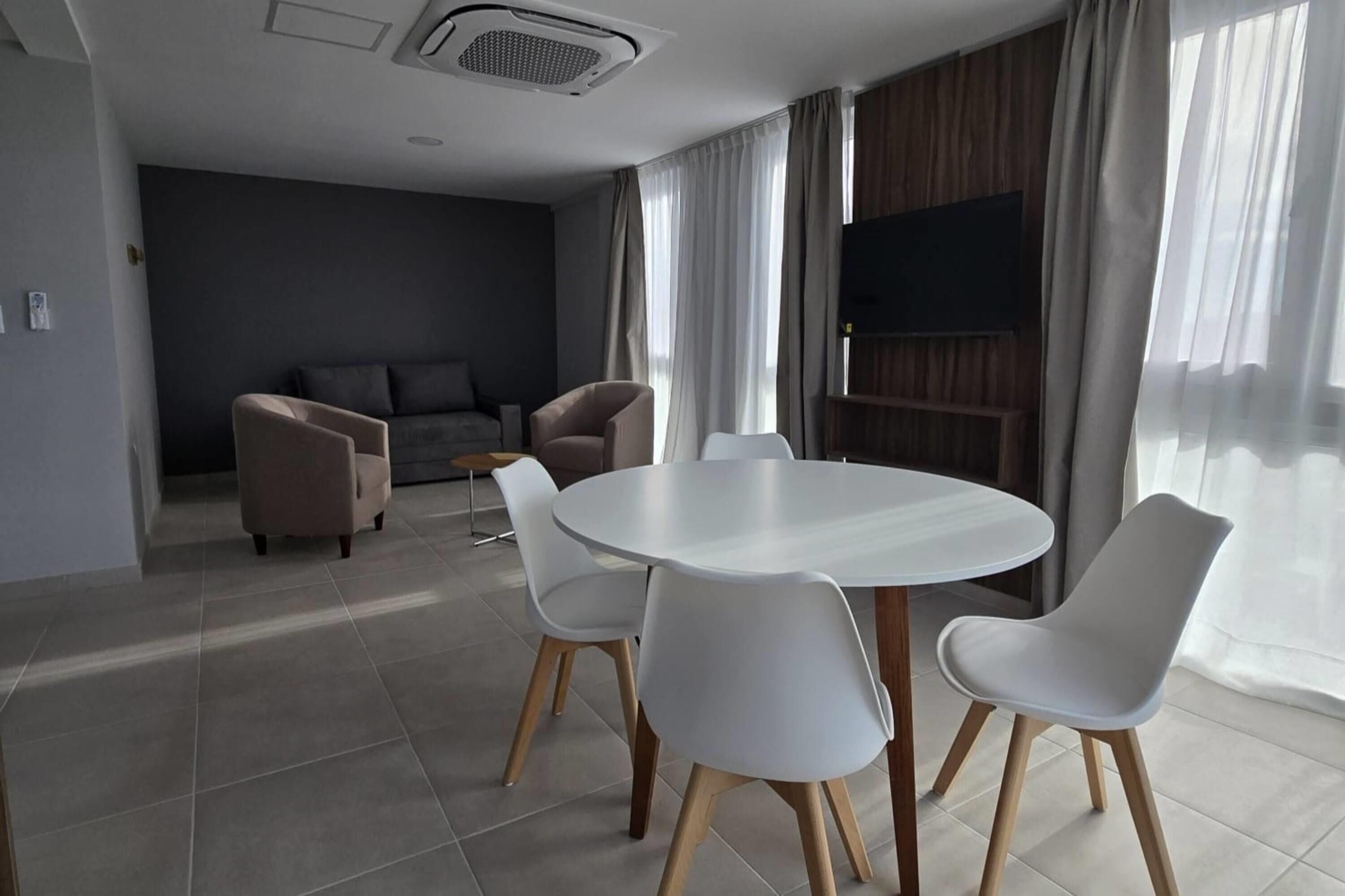 A modern living room with a round dining table, white chairs, a sofa, and a TV mounted on the wall.