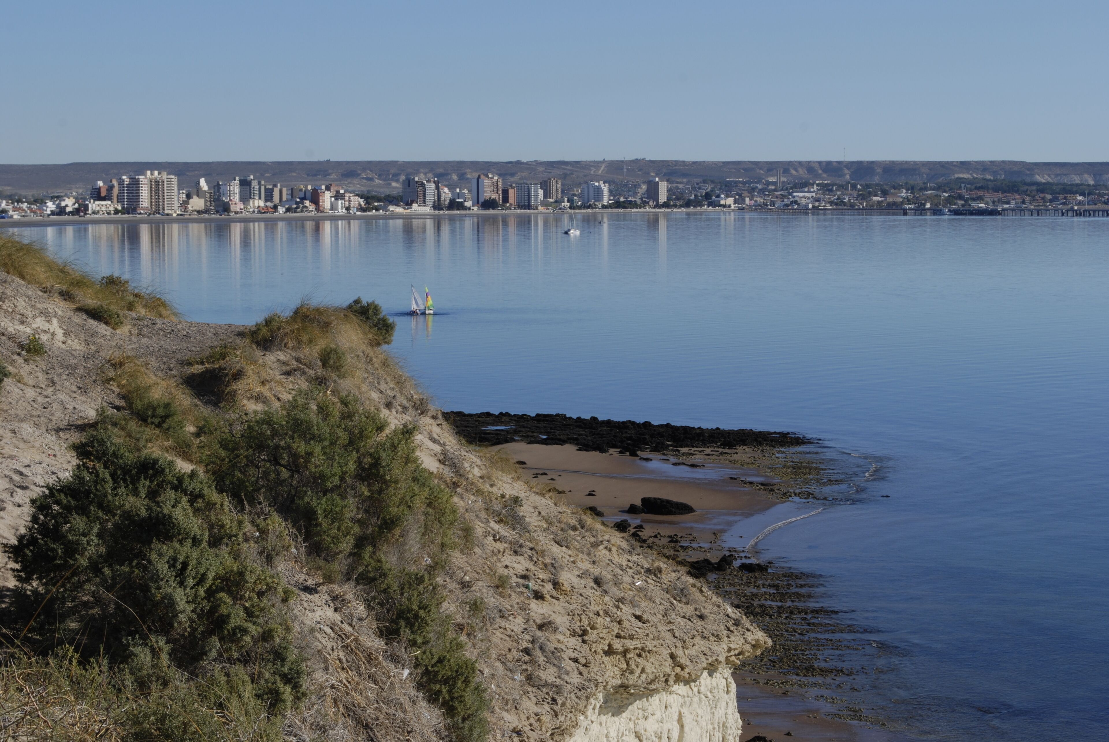 A coastal cityscape with a calm body of water, sandy beach, and a distant urban skyline.