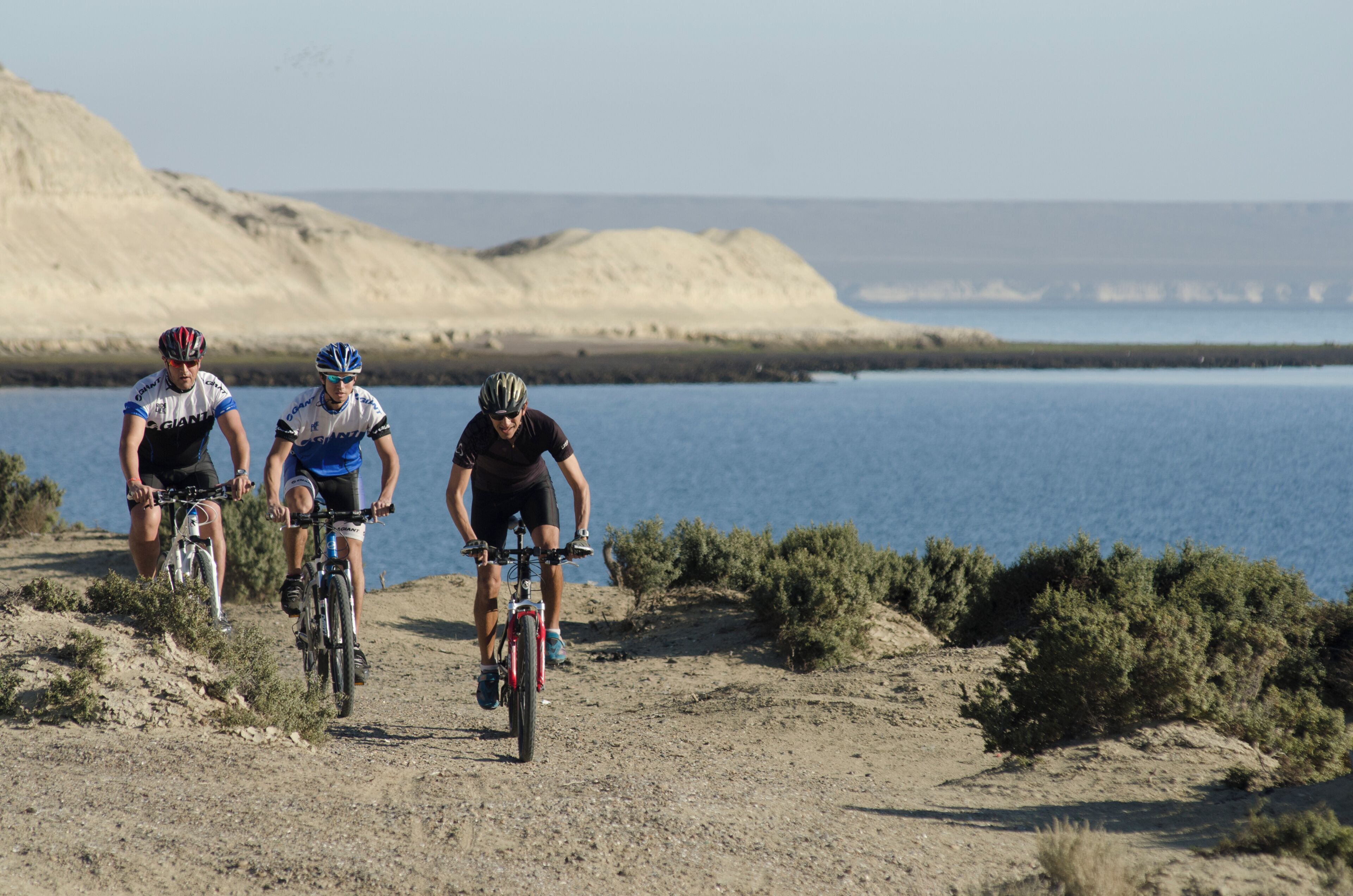 Três ciclistas numa trilha de terra com um corpo de água e colinas ao fundo.