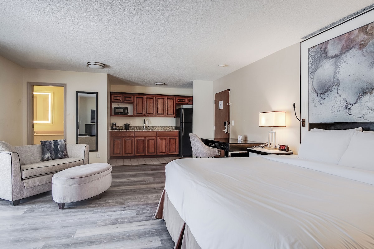 A hotel room with a large bed, a sofa, a small table, a chair, a desk, a lamp, and a kitchen area in the background.